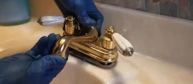 Blue gloved hands work on a brass two handle bathroom sink faucet