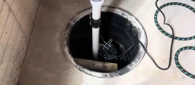 Round black sump pit in a basement floor with a white discharge pipe and pump cord